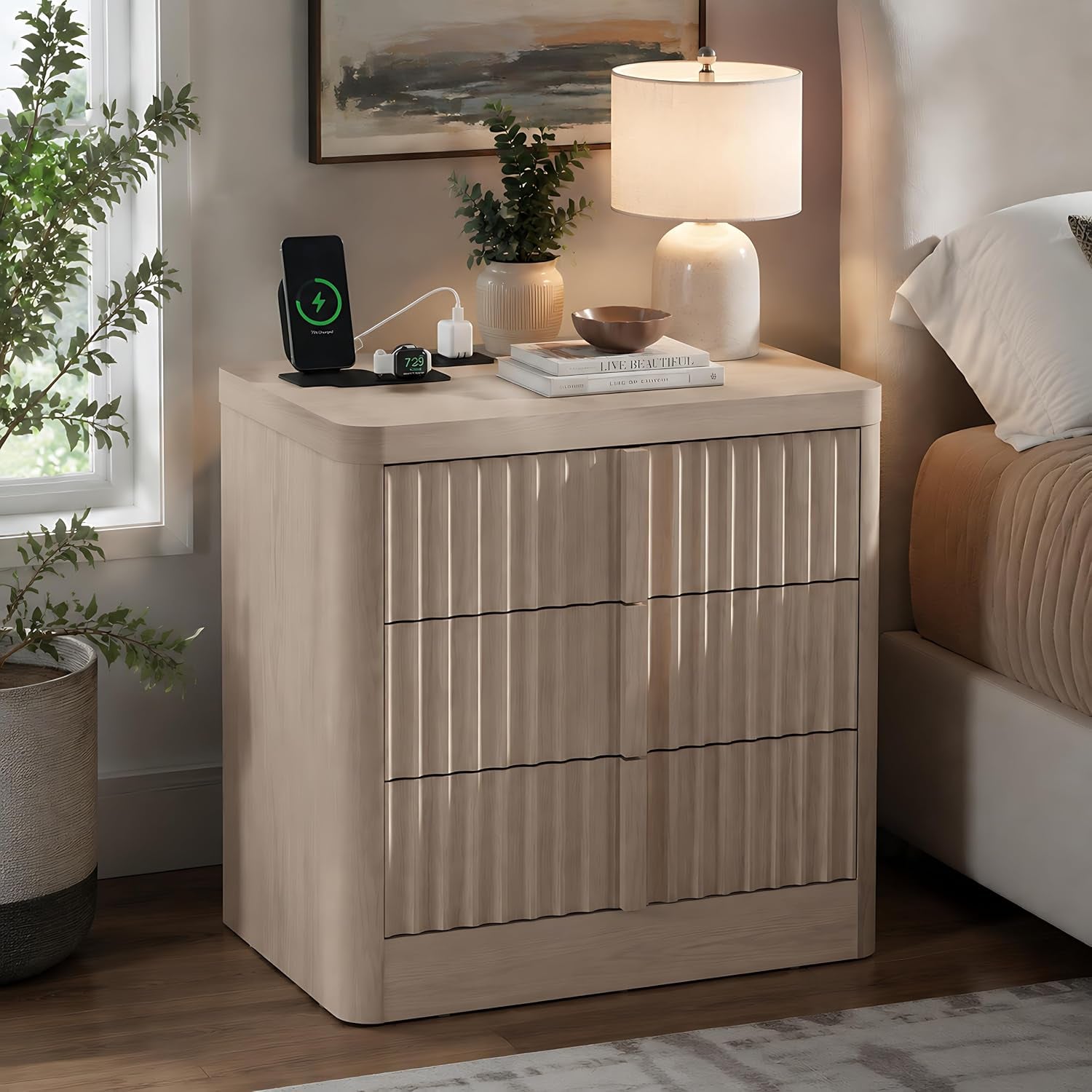 Light beige fluted nightstand beside bed with lamp, phone charging station and plant on top.