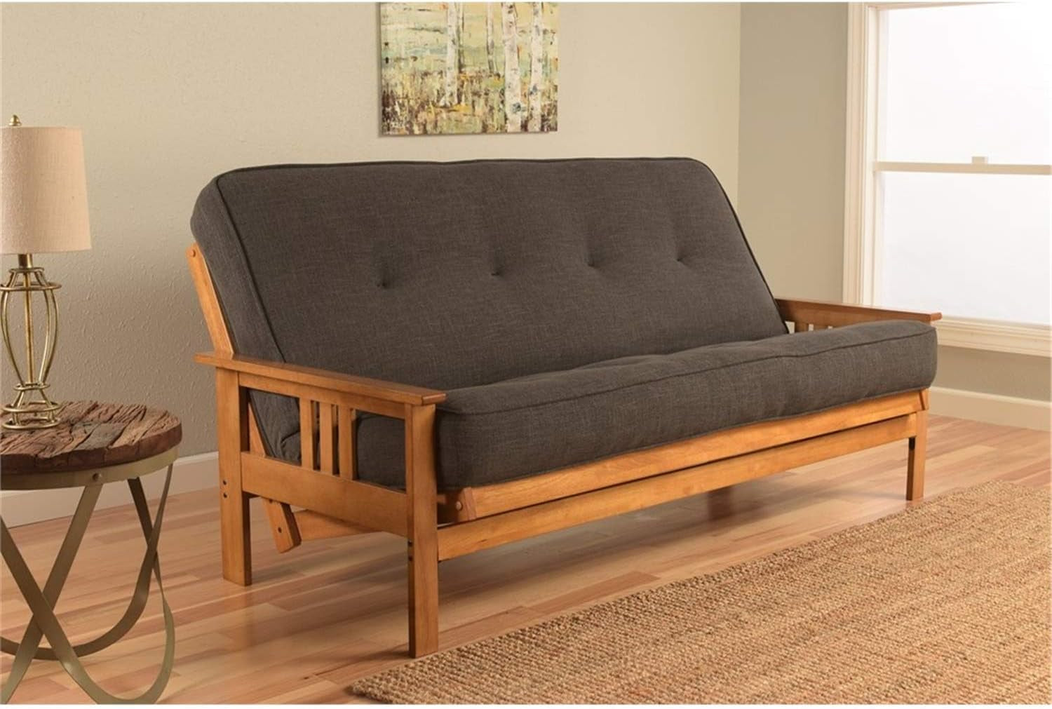 Living-room view of the futon in sofa position with a dark tufted mattress, side table lamp and framed art above.