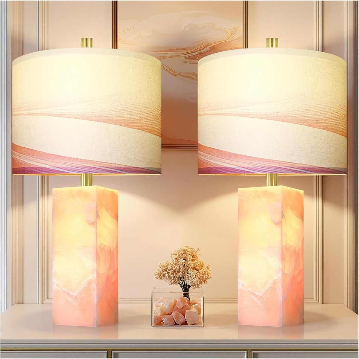 Two lit alabaster column lamps with pink-banded drum shades flanking a clear box of pale stones and dried flowers on a