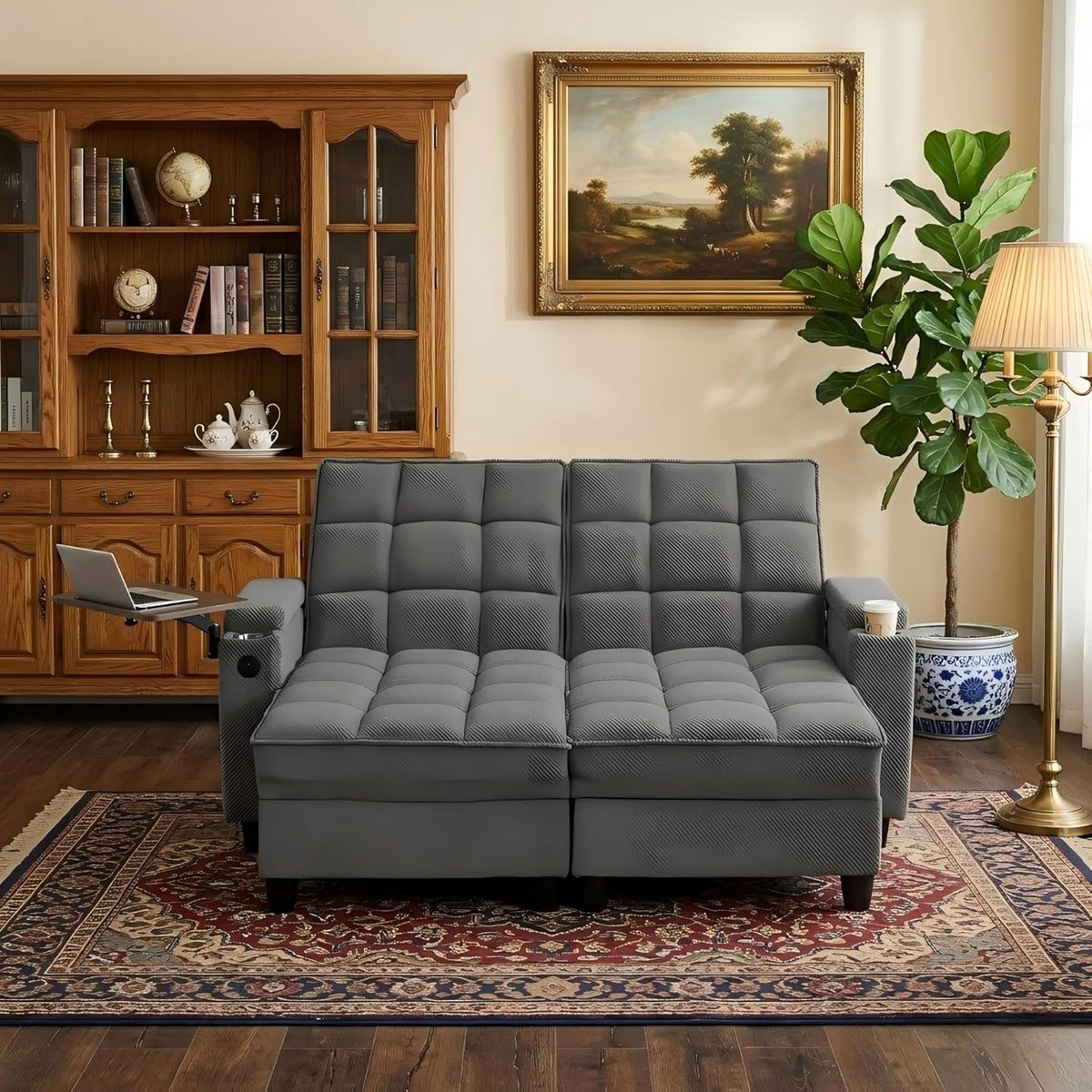 Gray tufted chaise sleeper fronted by patterned rug, laptop tray extended left, potted fiddle-leaf and framed landscape above