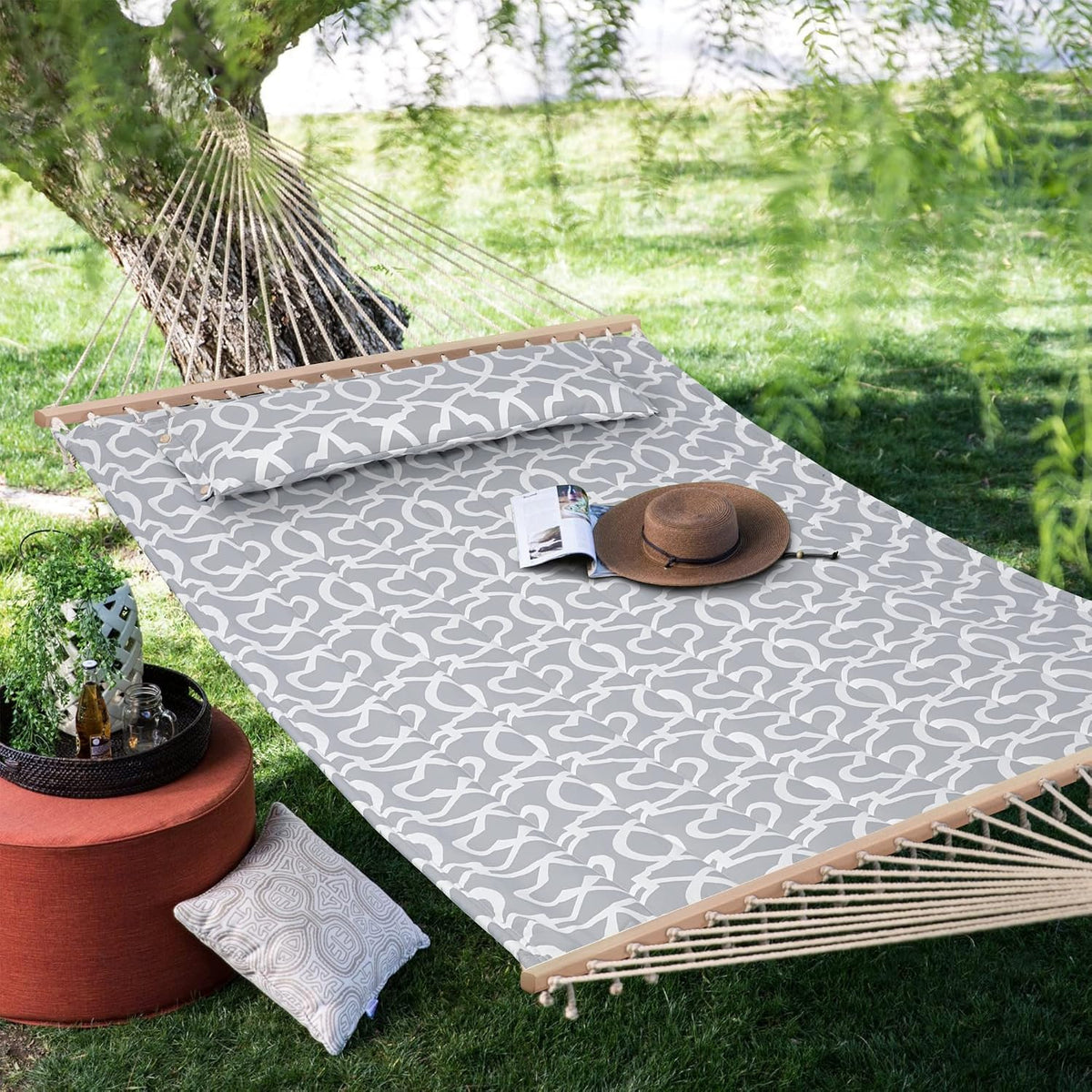 Grey patterned hammock set under a tree with a hat, open book and ottoman beside it