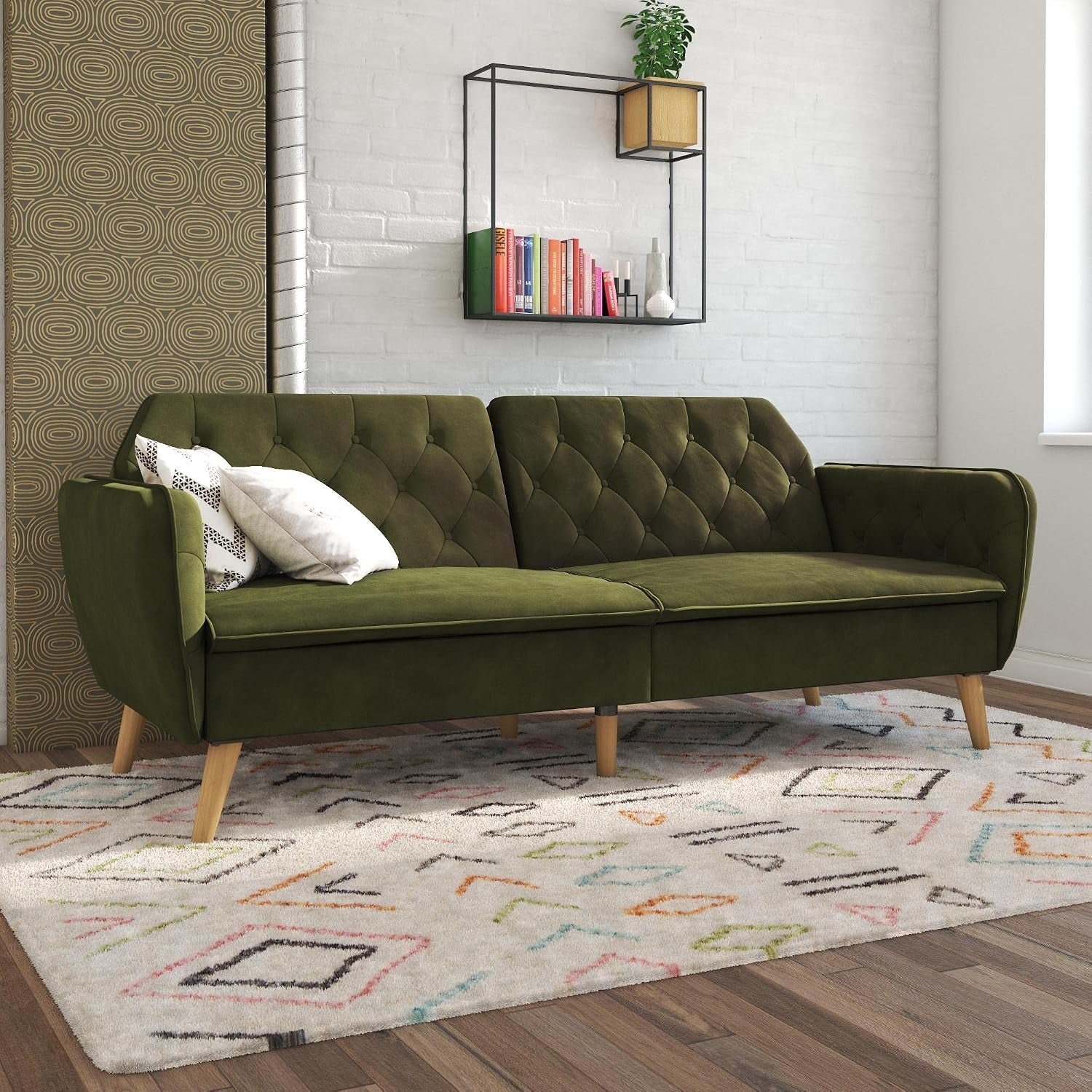 Green tufted futon sofa styled in a living room with patterned rug, pillows and wall shelving