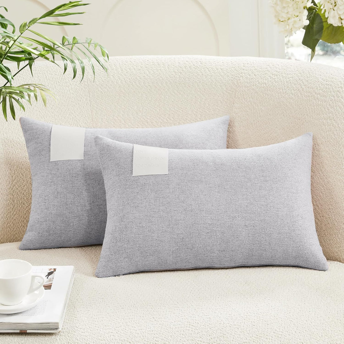 Two light gray rectangular lumbar pillow covers on a textured cream sofa with a plant and cup nearby.