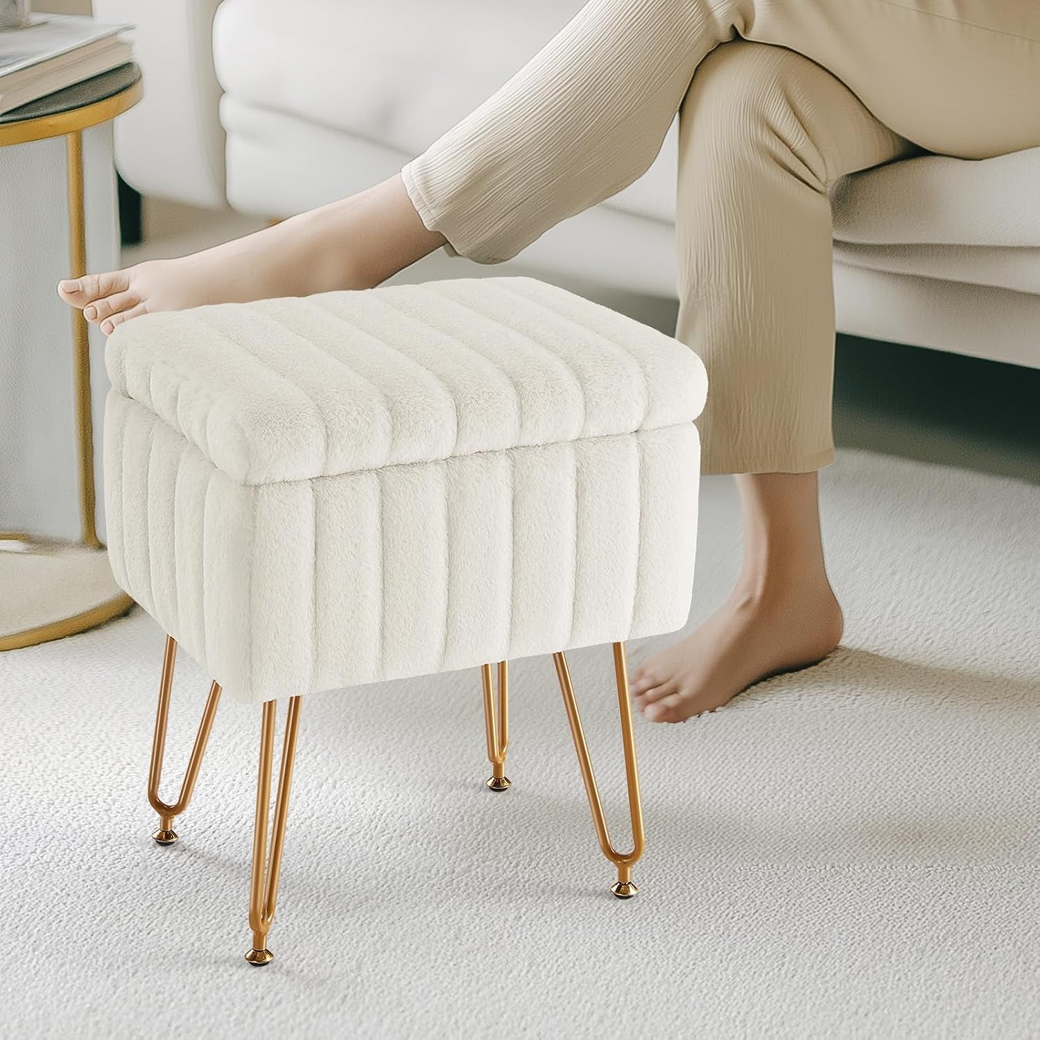 Cream faux-fur storage ottoman with vertical tufting and gold metal hairpin legs next to a sofa.