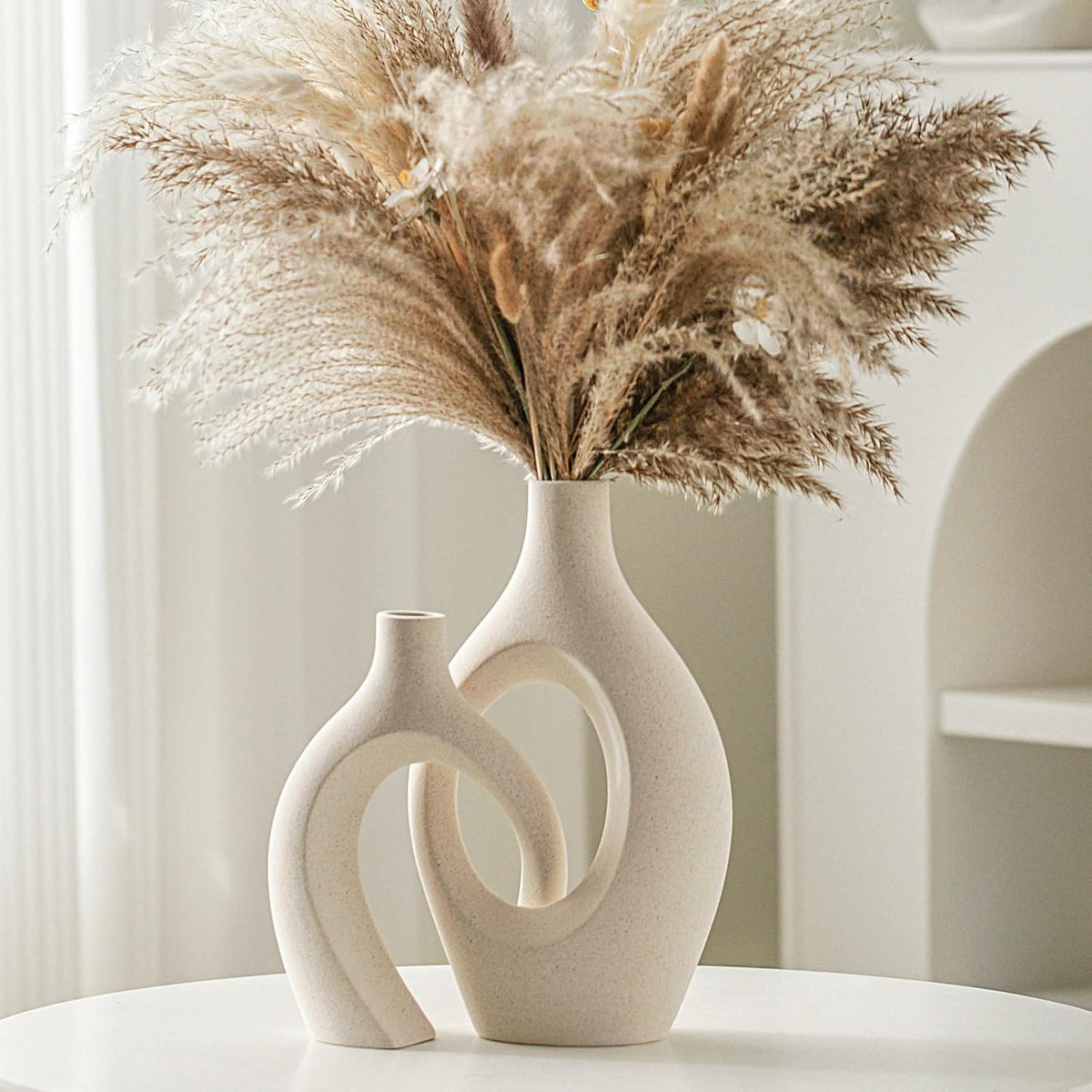 Vertical close-up of two interlocking beige ceramic vases filled with pampas grass, arch niche in background.