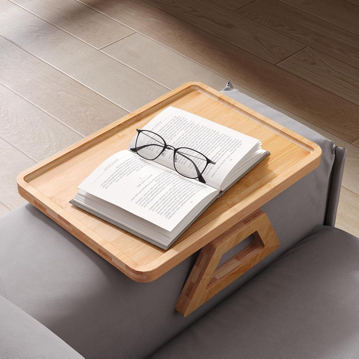 Angled top view of bamboo tray on a sofa arm with an open book and glasses resting on the tray