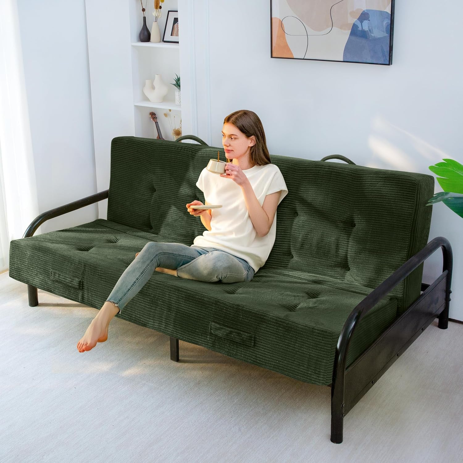 Dark green corded futon mattress set up as sofa on a black metal futon frame with a person sitting and holding a mug