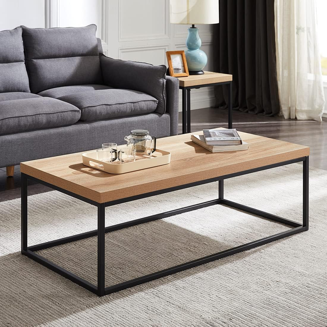 Light wood rectangular coffee table with black open metal frame, tray and glassware on top in a living room.
