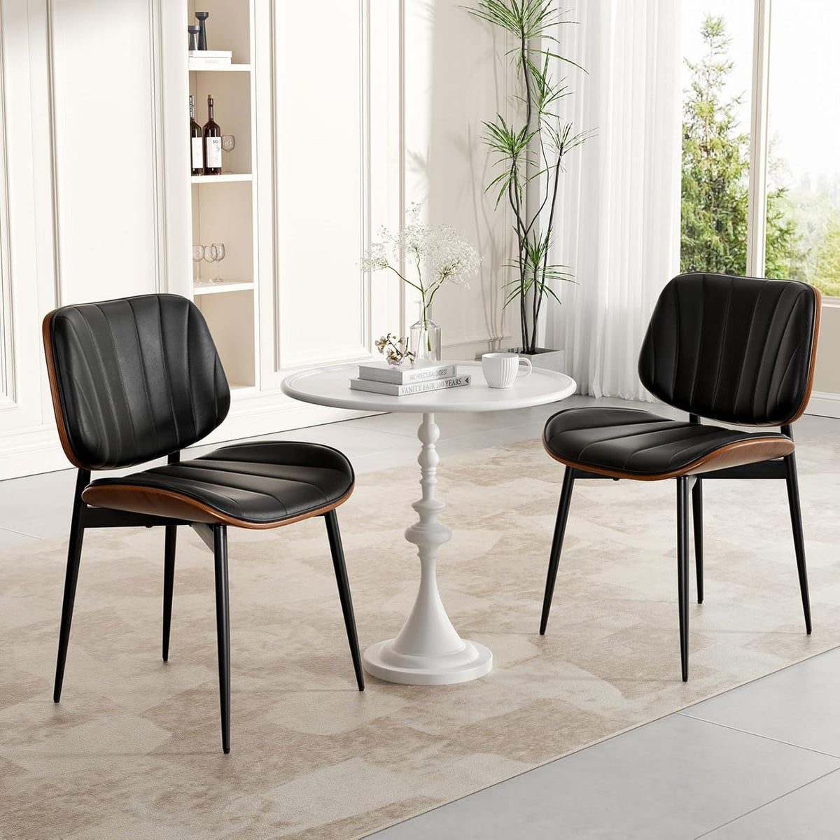 Two black padded chairs arranged around a small round white table in a bright dining nook with plants and paneled walls.