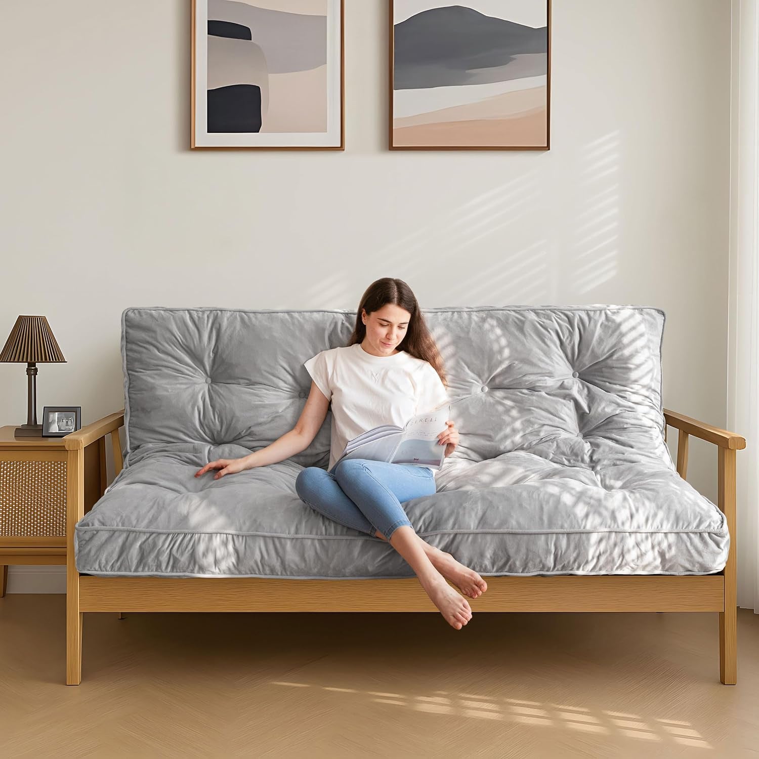 Light gray tufted futon mattress on a wood sofa frame with a woman seated and reading
