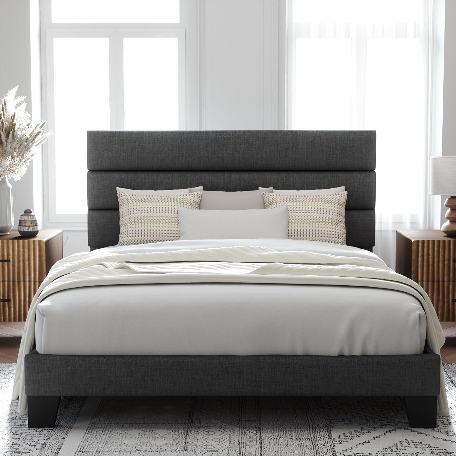 Front room view of a dark gray upholstered bed with a three-panel headboard, white bedding and black feet.