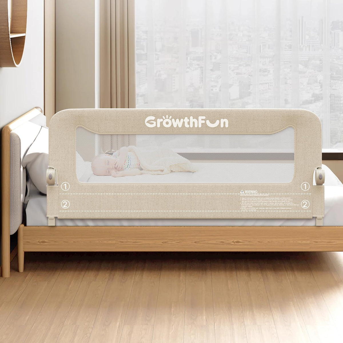 Front view of a beige toddler bed rail installed on a low wooden bed with a sleeping baby visible through mesh