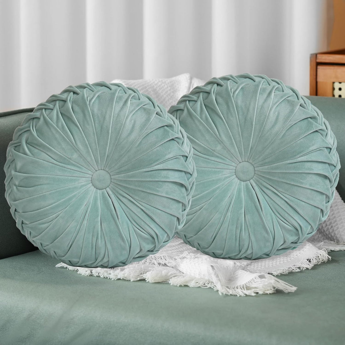 Two round ruched pillows in muted teal placed on a sofa with a fringed white throw.
