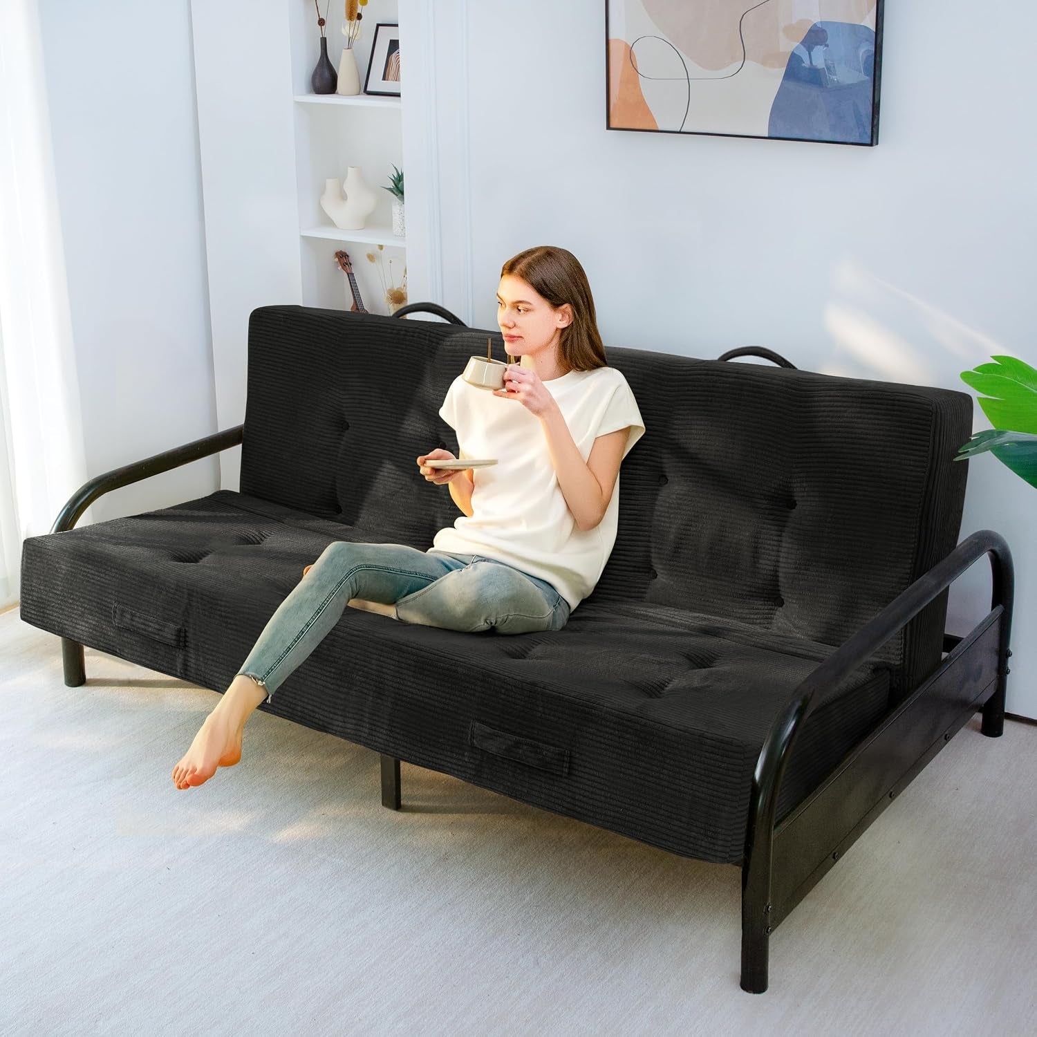 Woman sitting on a black corded futon mattress with tufted buttons on a black metal frame, sipping from a cup.