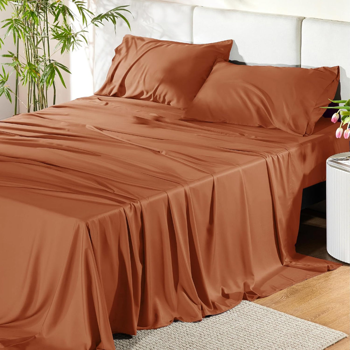 Warm terracotta-colored sheets and two pillows on a bed beside a small round side table and plant.