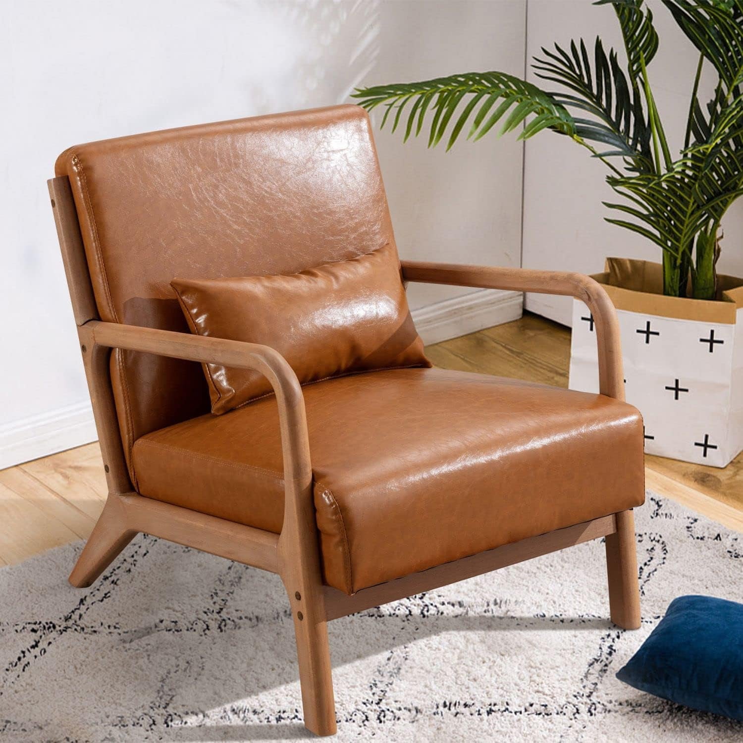 Angled close view of the chair showing leather seat, wooden arms and tapered rear legs with plant in background.