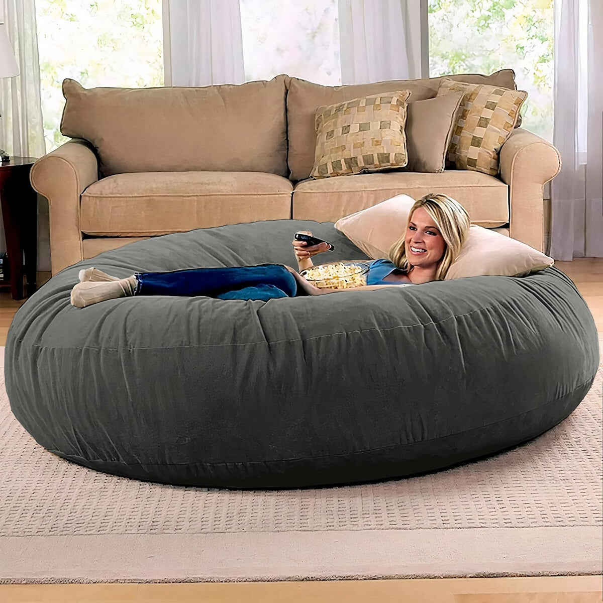 Charcoal-grey round giant bean bag bed flat configuration with a person reclining in front of a beige sofa