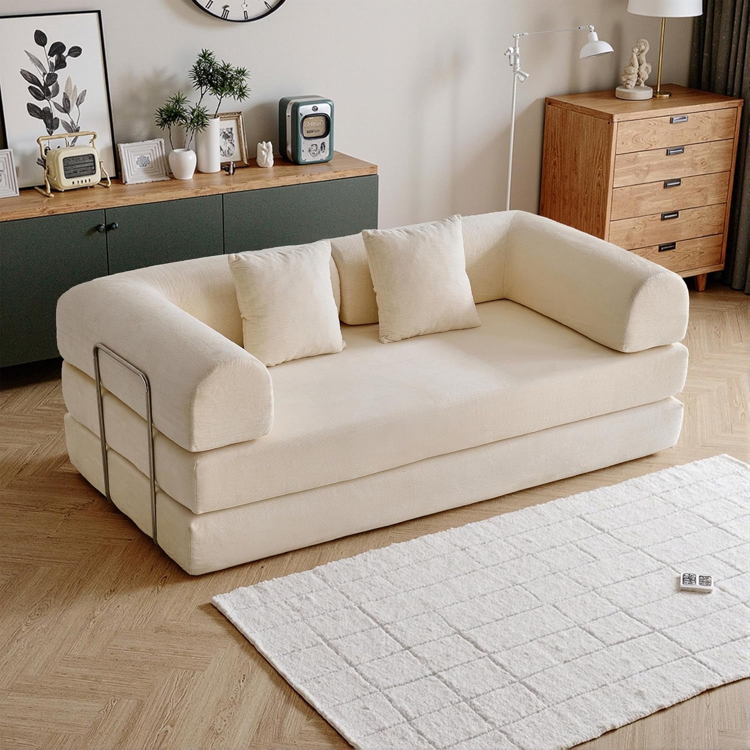 Cream-colored folding sofa with rounded arms, three cushions and metal side support in a bright room.