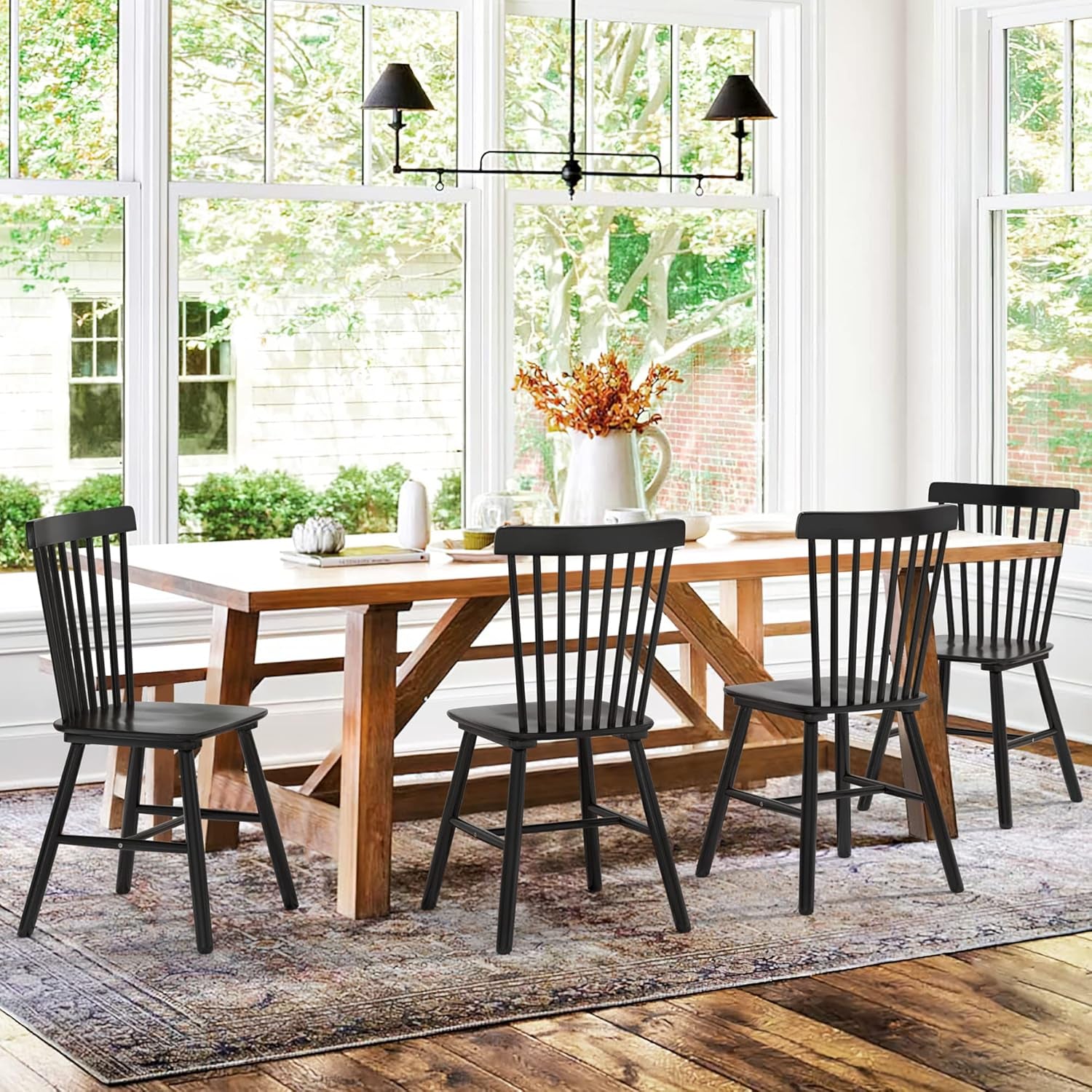 Dining area with four black spindle-back chairs arranged around a farmhouse-style wooden table by large windows.