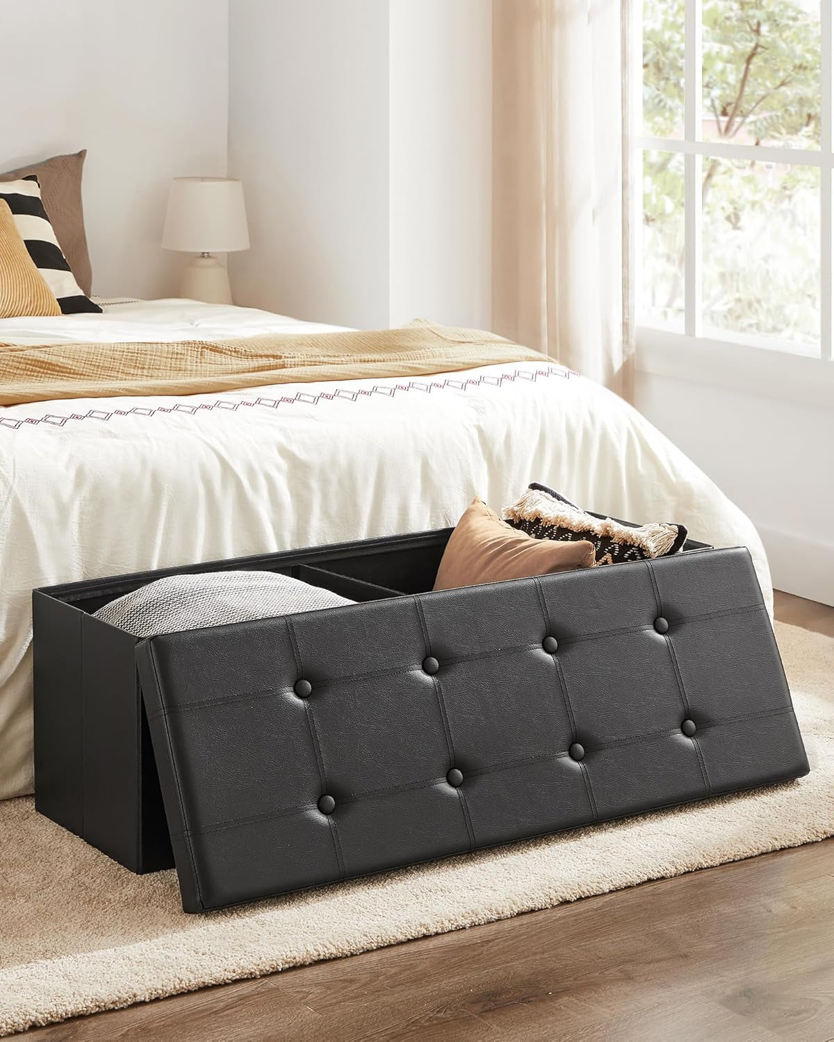 Black storage ottoman open with lid removed showing internal divided storage beside a bed