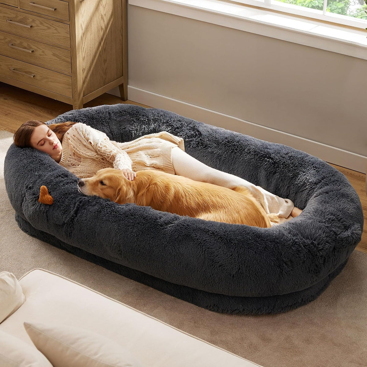 Woman lying inside an oversized charcoal-gray plush dog bed with a golden retriever stretched beside her.