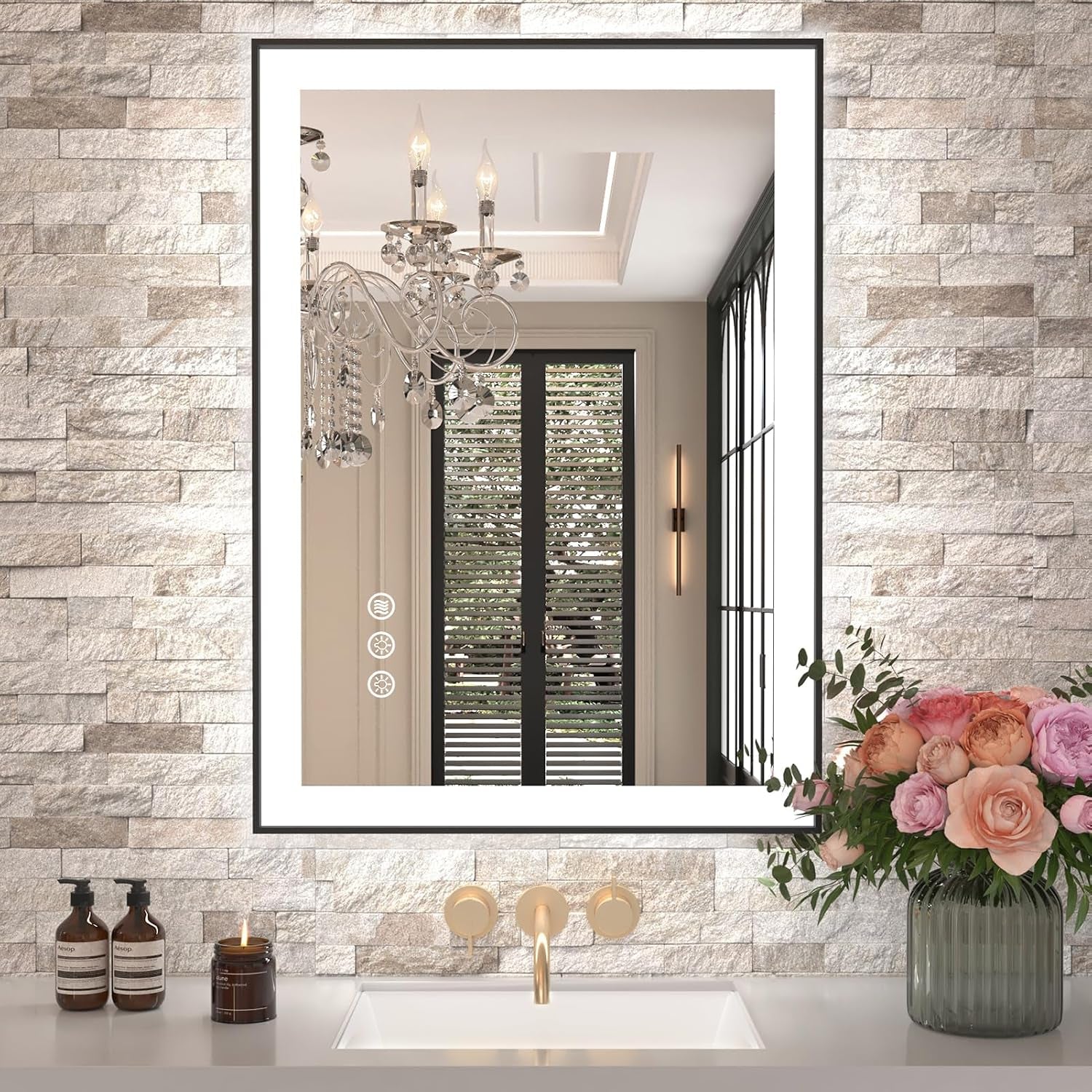 Tall rectangular mirror with illuminated inner border mounted on textured stone wall above a sink with flowers on the