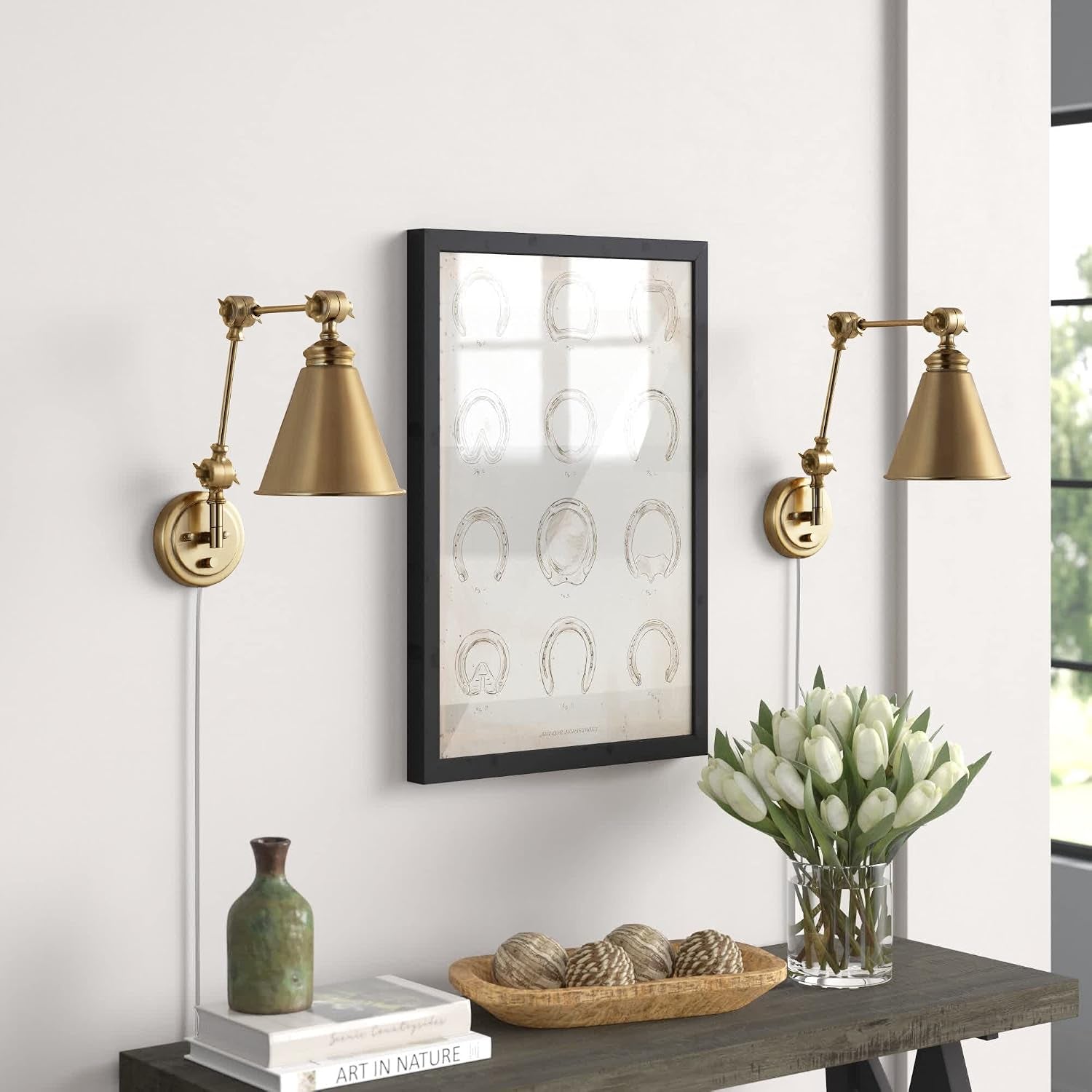 Brass swing-arm wall sconces mounted above a console table with visible plug-in cords and framed art.
