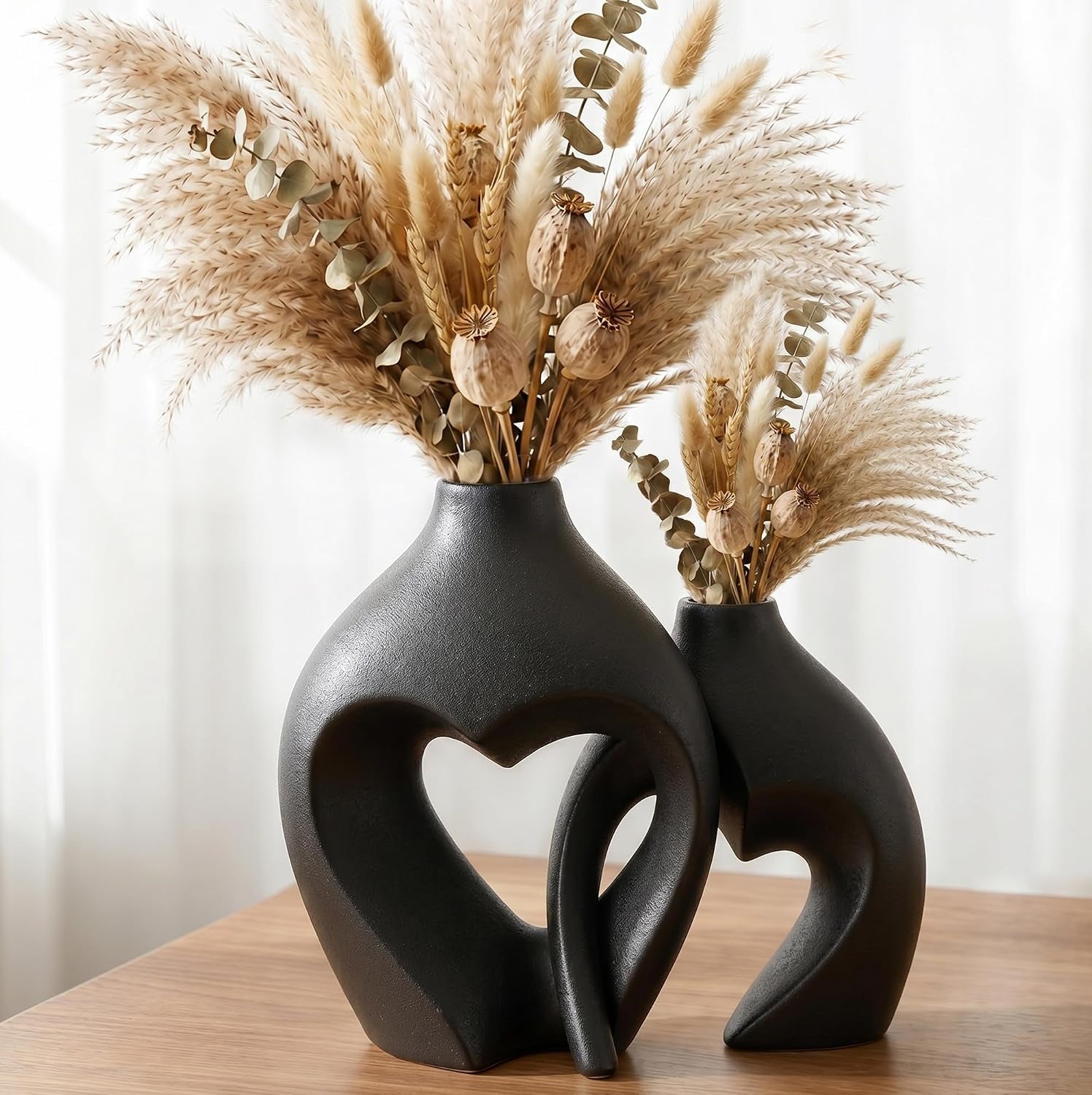 Two dark matte heart vases in charcoal on a wood table filled with pampas and dried stems.