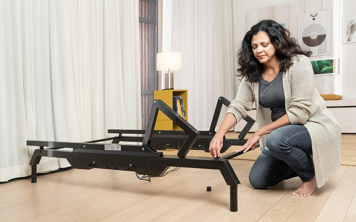 Woman assembling black adjustable base on hardwood floor, kneeling while adjusting frame legs.