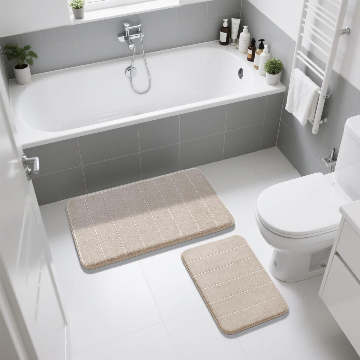 Overhead view of two beige bath mats placed on a white tiled bathroom floor beside a tub and toilet.