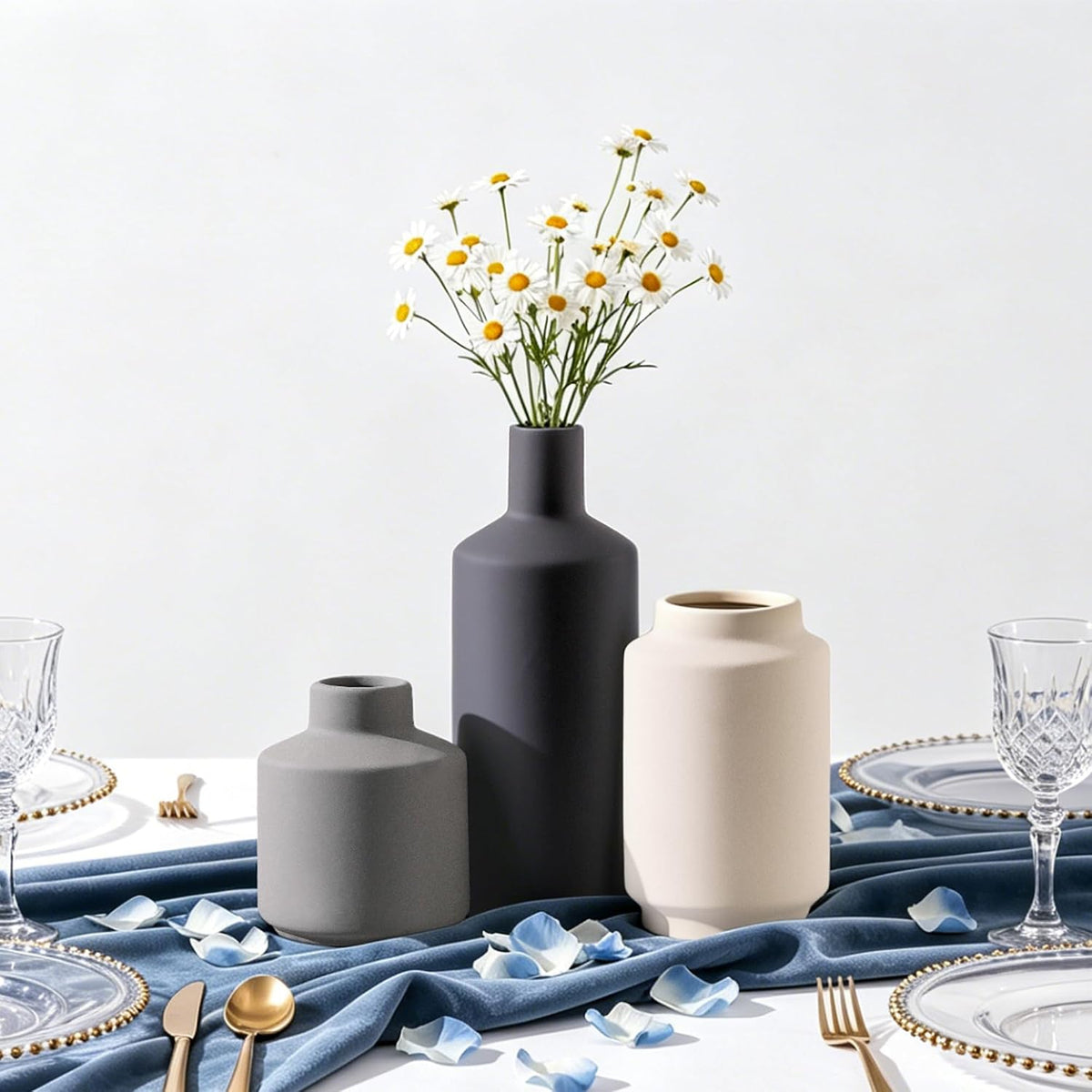 Three matte vases on a draped blue cloth with scattered petals and glassware, daisies in the tall vase