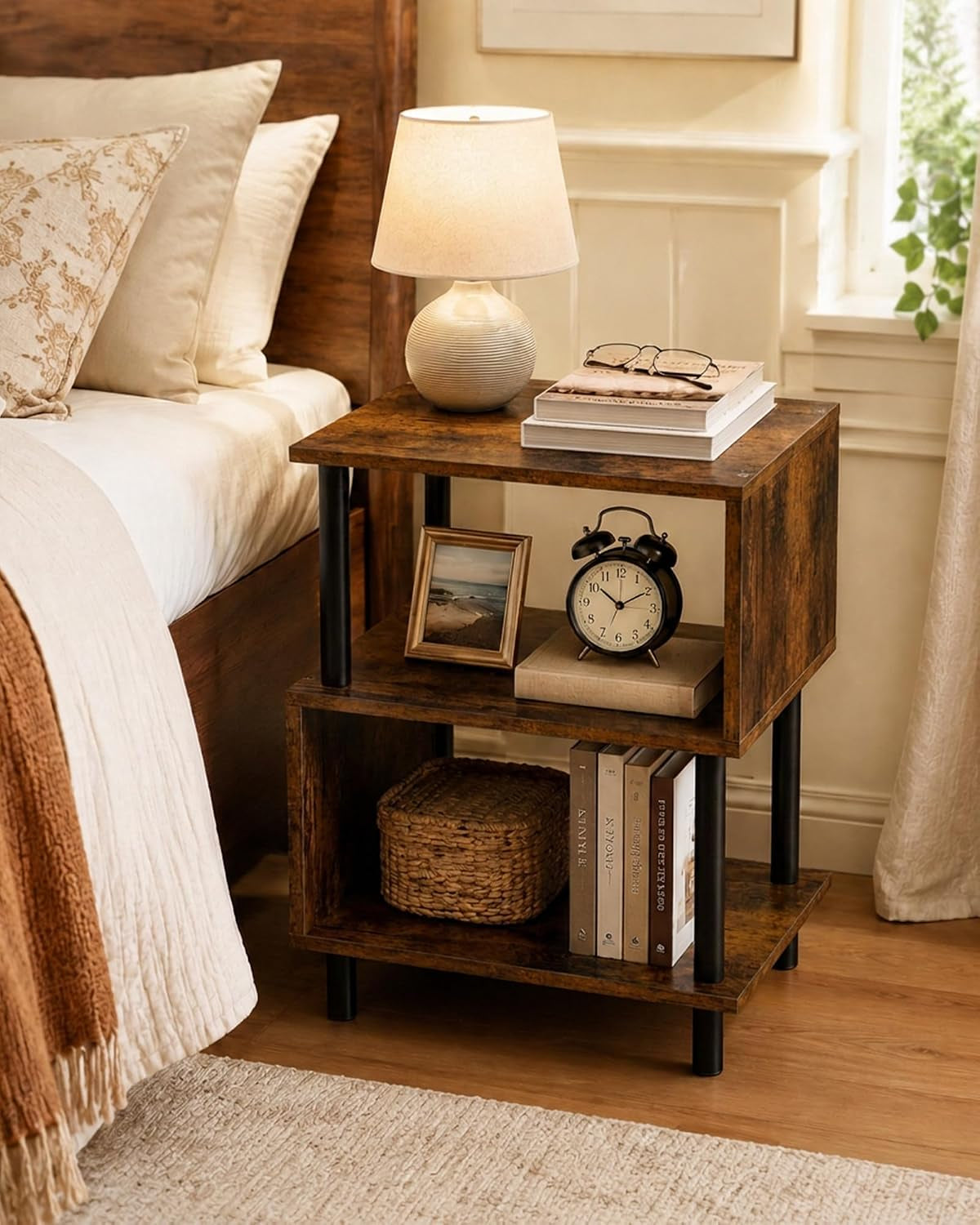 Rustic brown 3-tier side table used as a nightstand beside a bed with lamp and clock on display