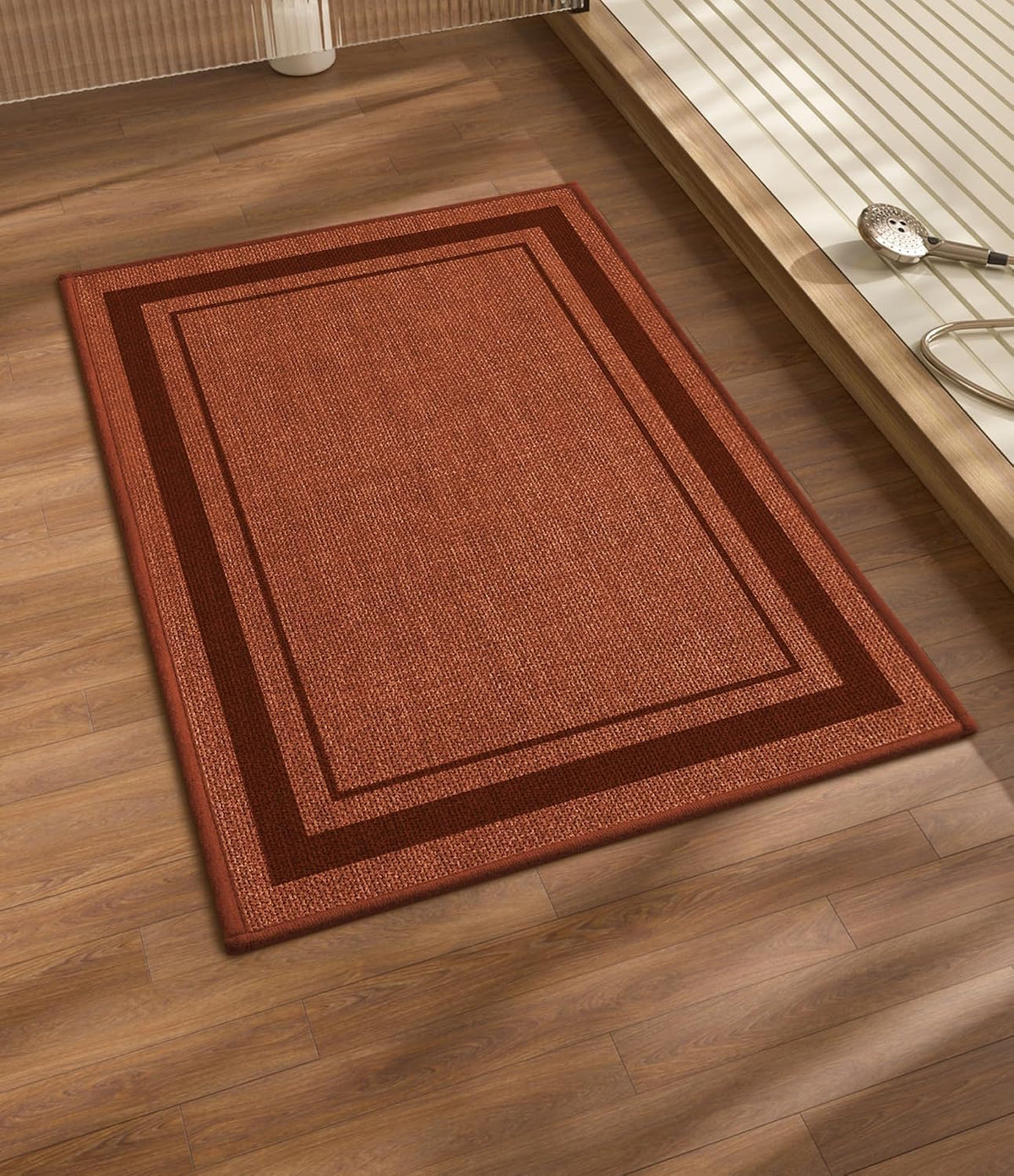 Rectangular rust-colored bath mat with double-border on wood floor beside a tub edge.