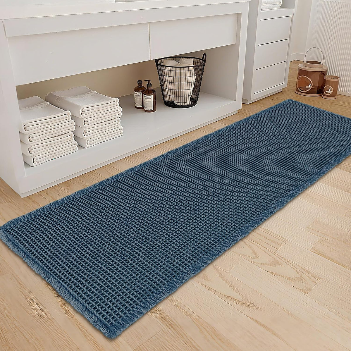 Waffle-pattern runner bath mat in muted teal blue on a light wood floor beside folded towels and basket.