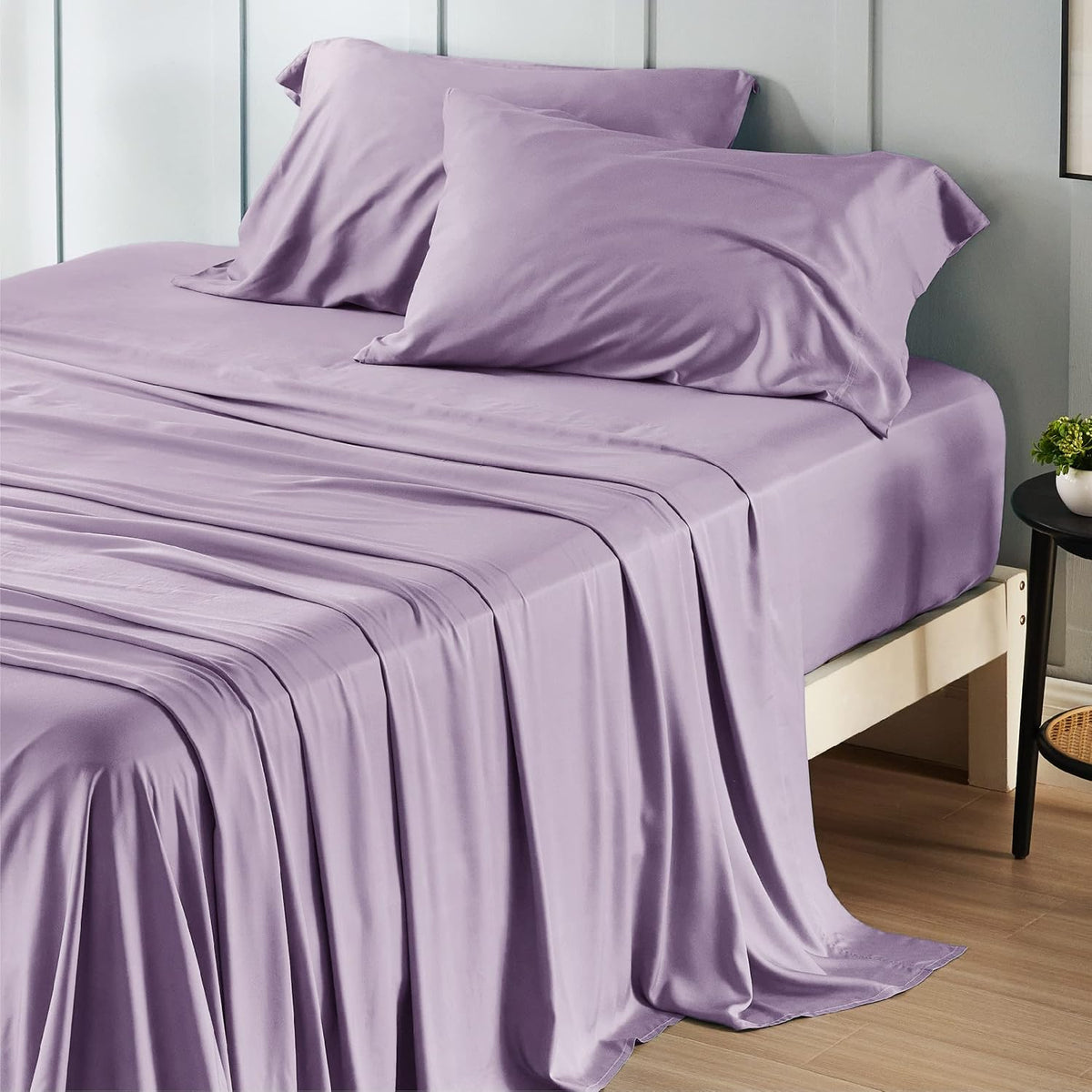 Side-on view of a bed dressed in lavender silky sheets with the sheet draped to the floor at the foot.