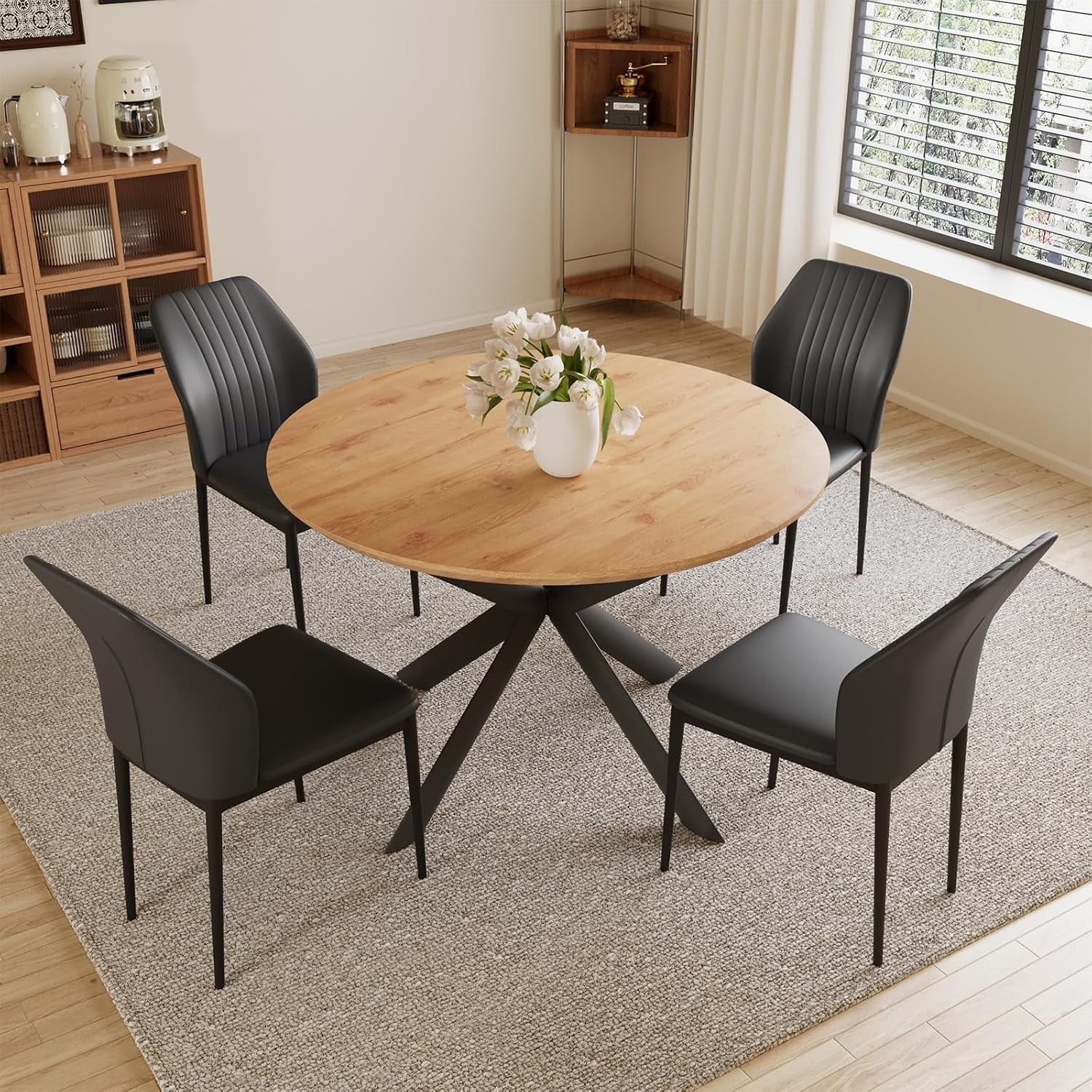 Round light-wood dining table with four black chairs placed on a neutral rug under a window.