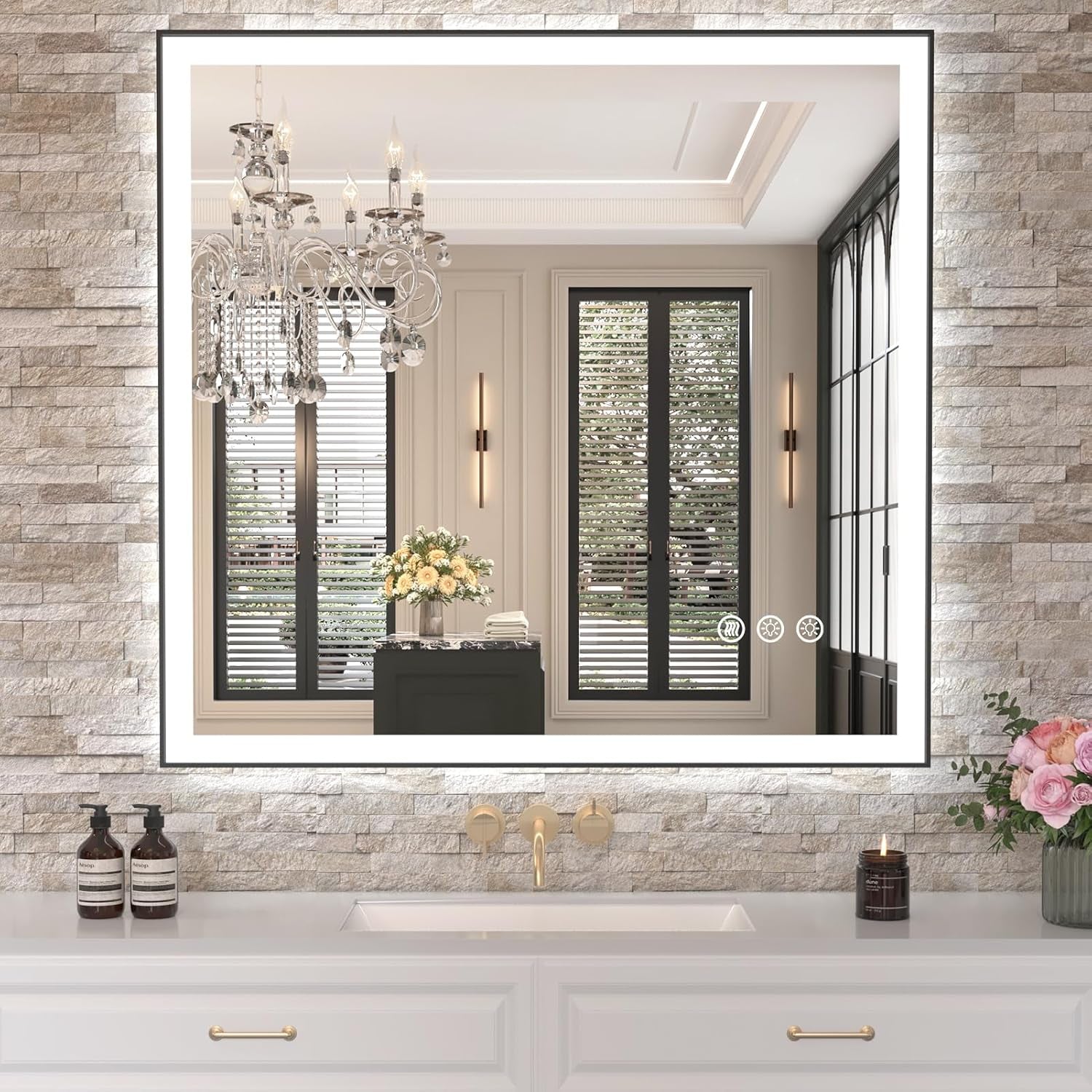 Square illuminated mirror on a textured light brick wall reflecting windows and a chandelier above a white vanity.
