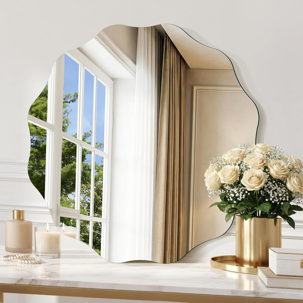 Frameless scalloped round mirror reflecting a window, styled on a marble vanity with roses and a candle