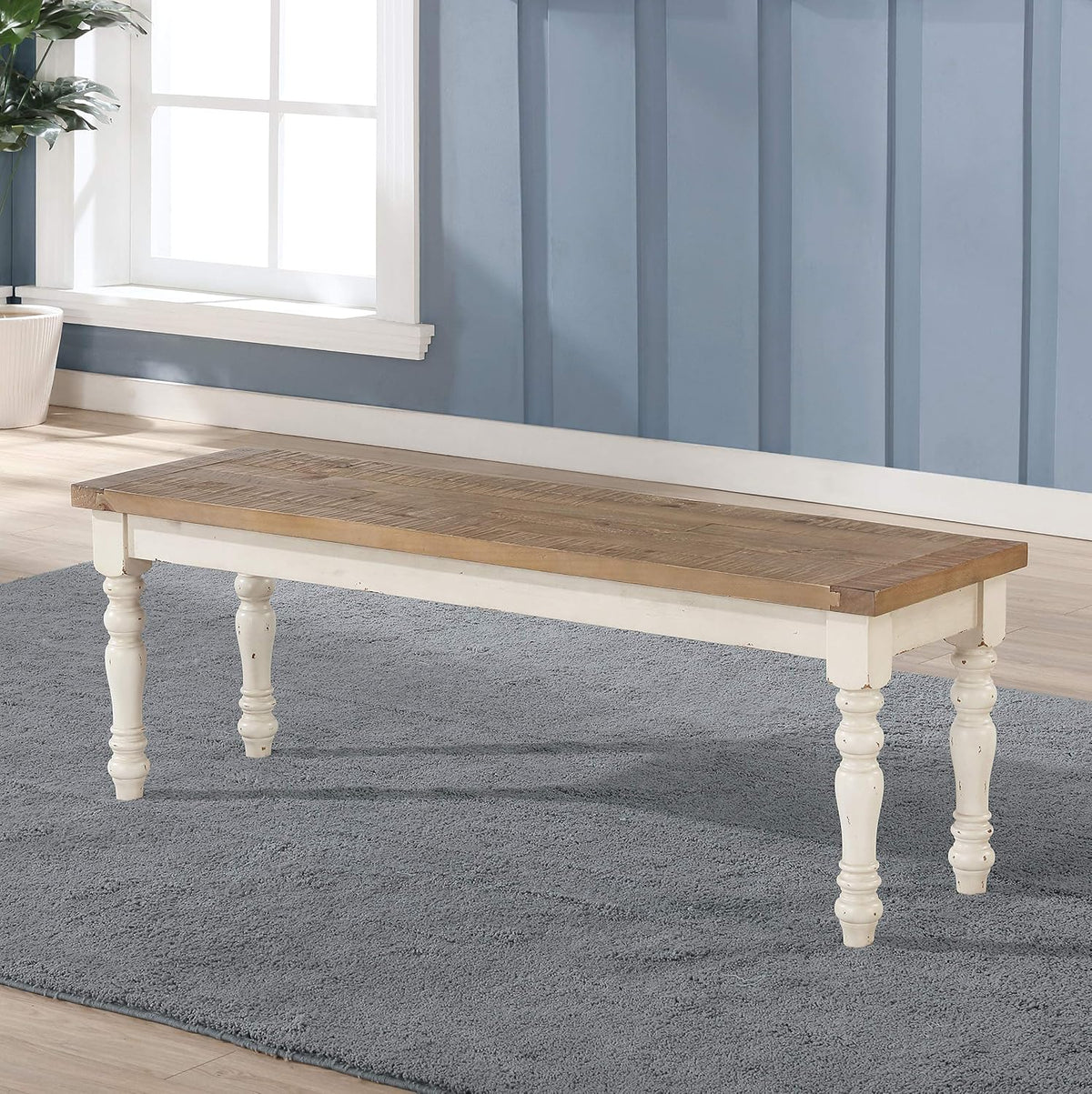 Three-quarter view of the bench on a gray rug beside a window, showing the wood top and distressed cream legs.