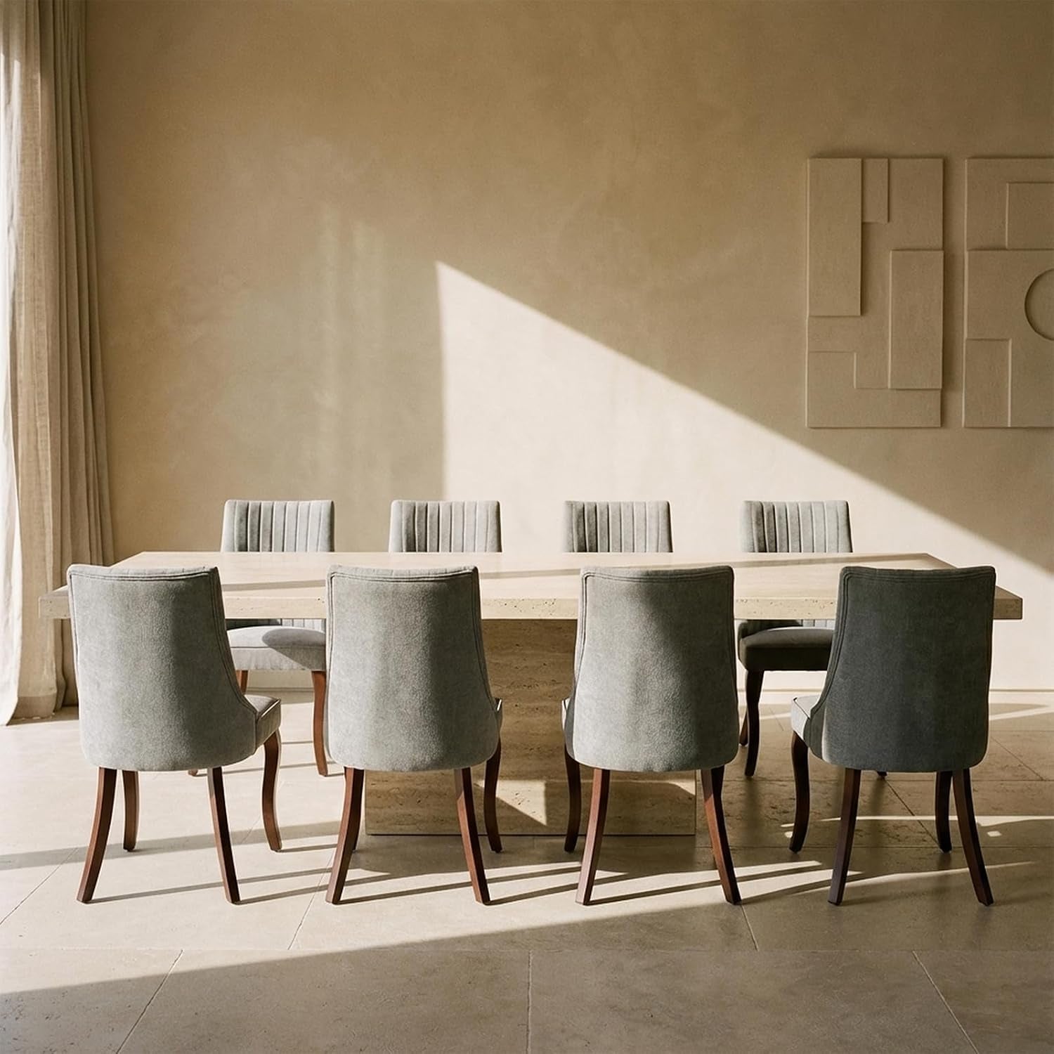 Back view of eight beige/grey upholstered dining chairs pushed to a long stone table under wall art.