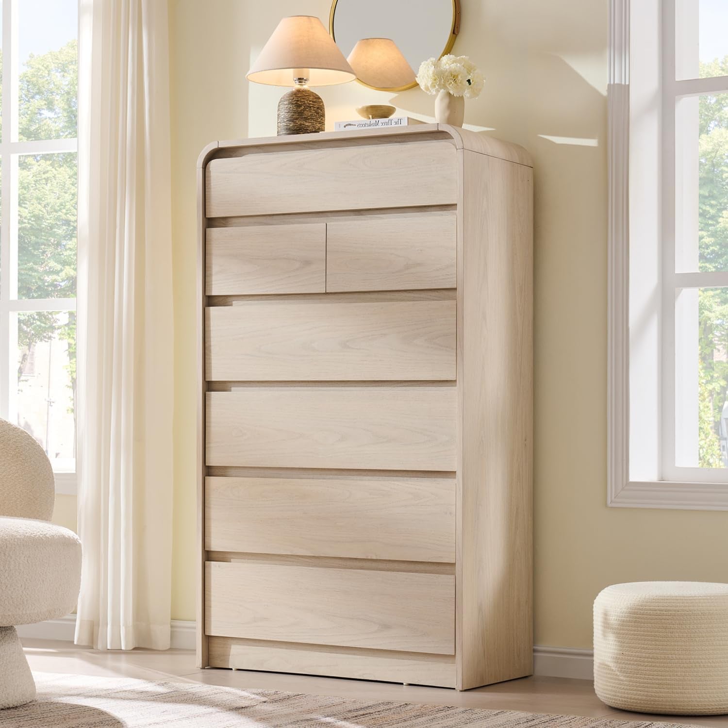 Tall natural oak dresser seen slightly from the left with a round mirror and lamp on top, pouf at right.