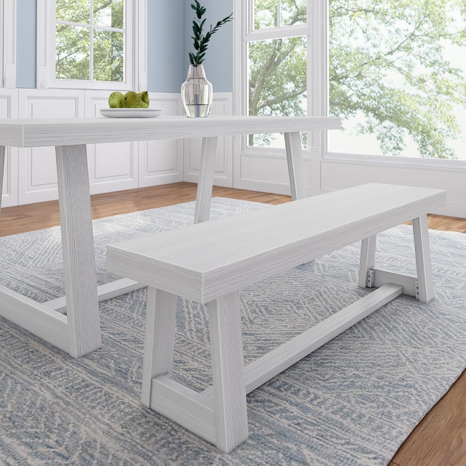 Dining table and matching bench in a bright whitewashed finish on a patterned rug by sunlit windows