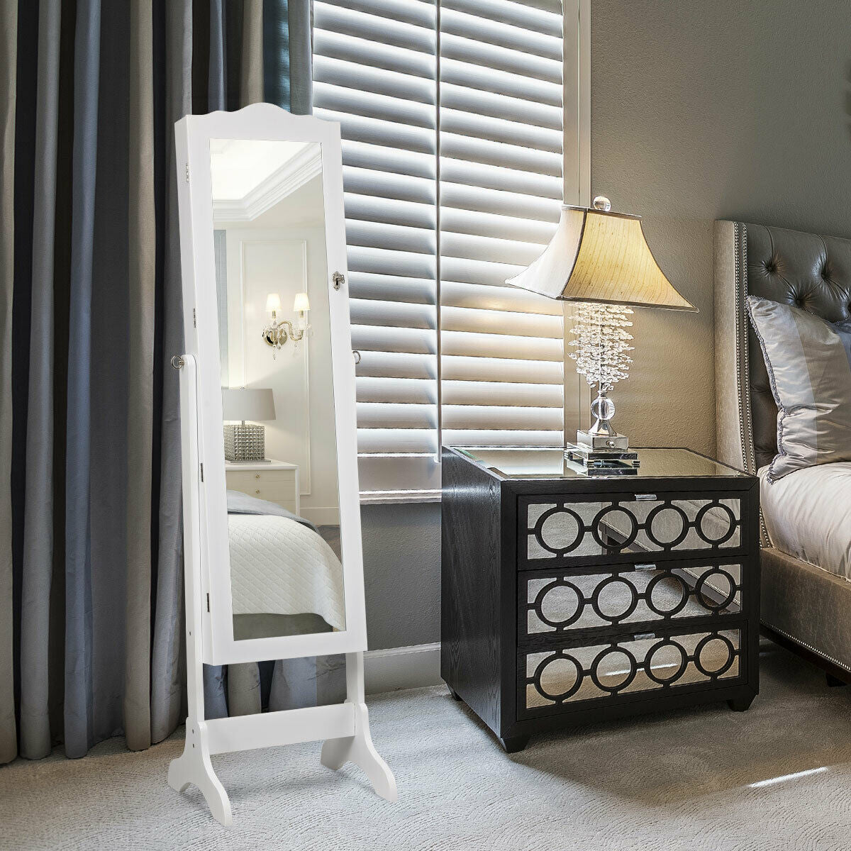 Closed white full-length mirror armoire standing beside a bed and window in a bedroom.