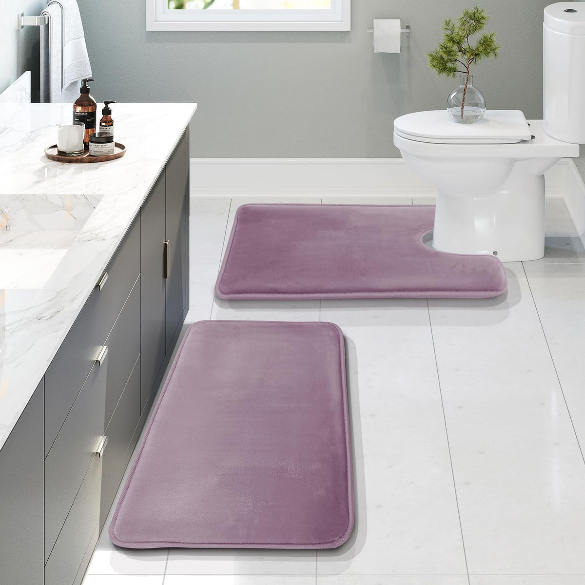 Staged modern bathroom with two mauve memory-foam rugs on glossy white tile beside a gray vanity and a toilet.