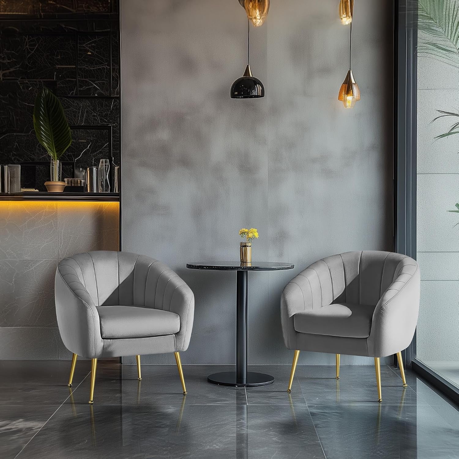 Two gray barrel chairs with gold legs facing a small round black table under hanging pendant lights in a modern space.