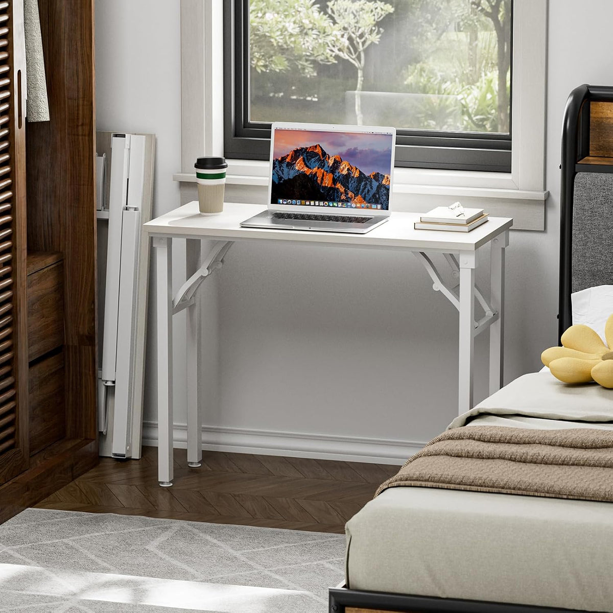 Desk placed under a window in a bedroom, styled with a laptop, coffee cup and stacked books