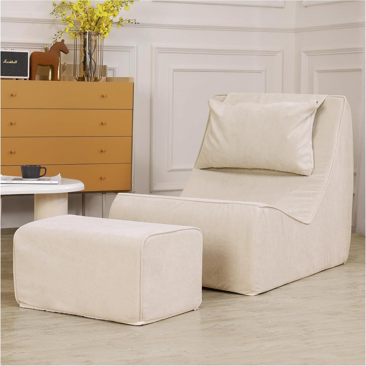 Cream-colored chaise and ottoman in a neutral room with a mustard chest and yellow flowers on top