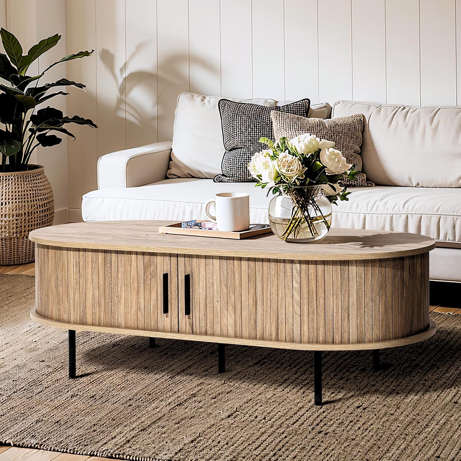 Oak-grain oval coffee table with sliding tambour doors and vase on top, angled front view in a bright living room