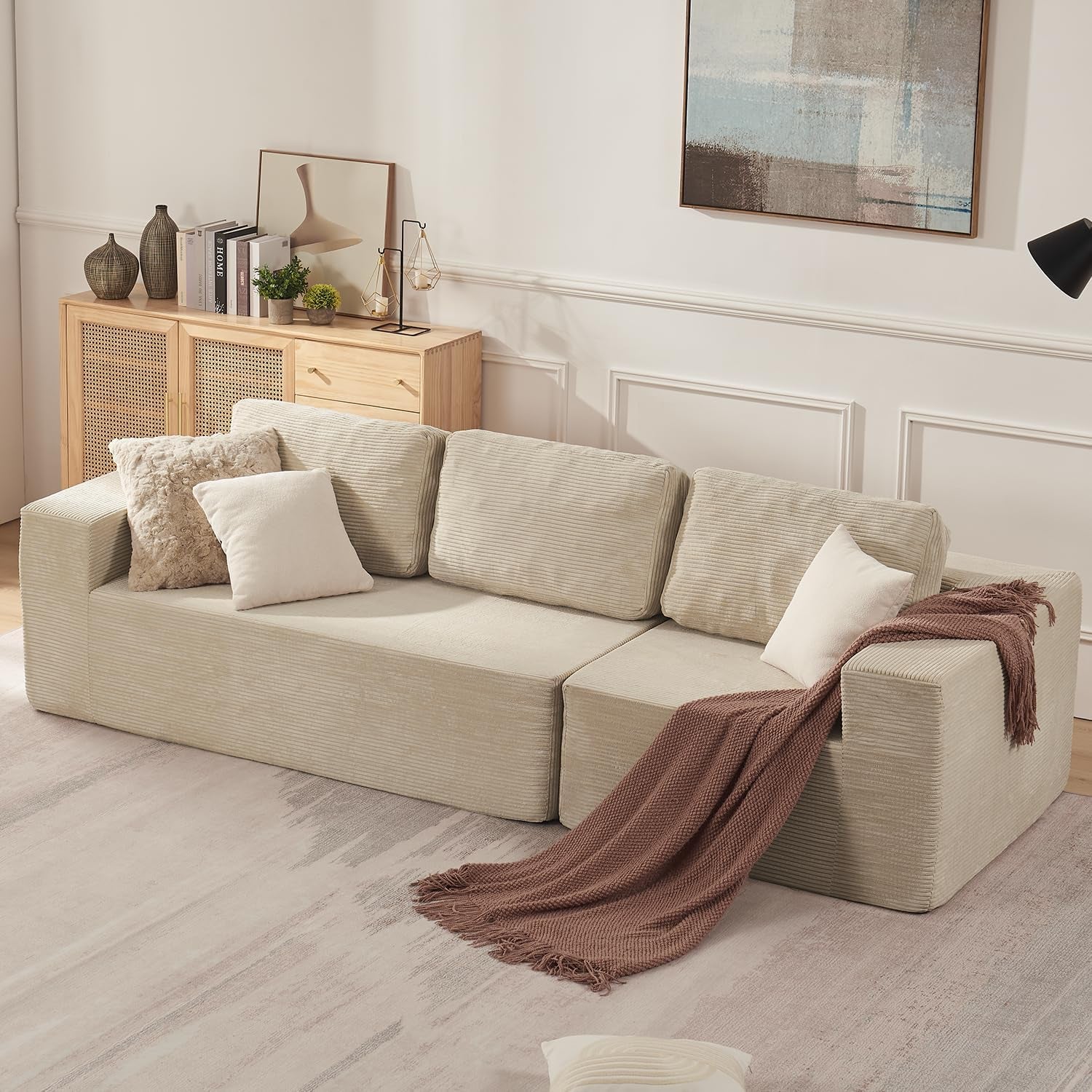 Light beige modular sofa with low arms, shown with textured pillows and a rust-colored throw on a rug.