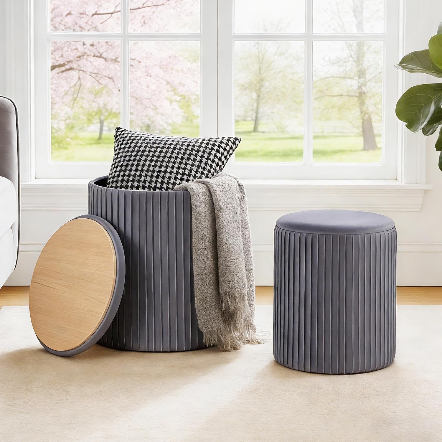 Gray round ottoman set staged by a bright window on a rug, larger ottoman open with pillow and blanket inside.